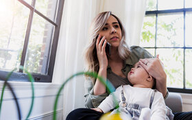 Eine junge Mutter hält die Stirn ihres Kindes und ruft ein Baby-Telefon für Rat an.