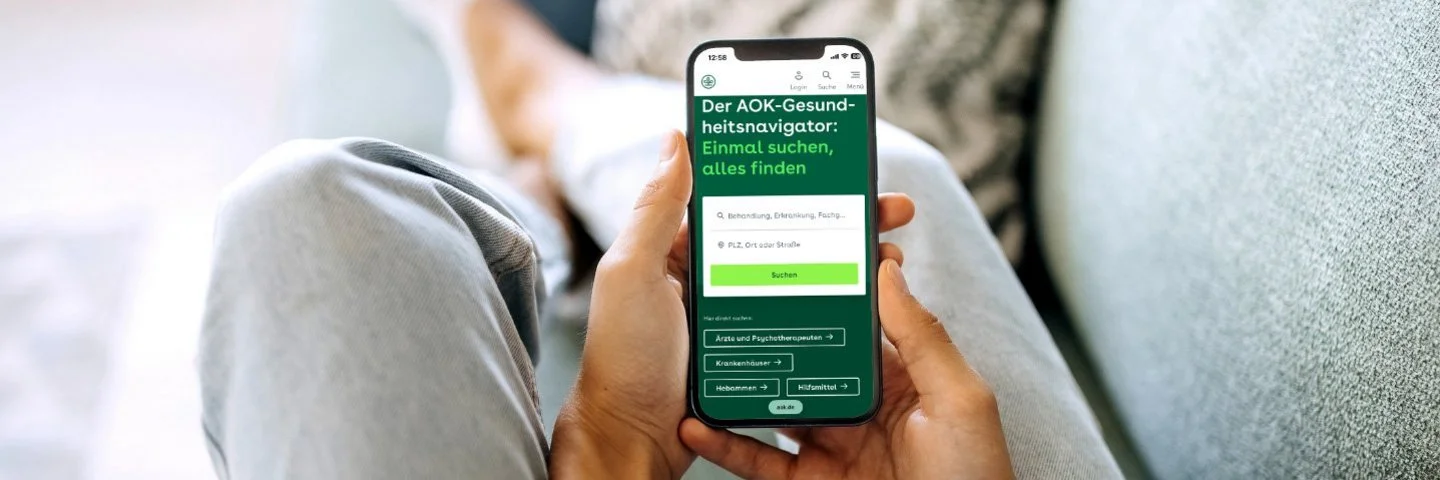 Eine nicht erkennbare Person sitzt auf einem Sofa und hält ein Smartphone in den Händen, im Display ist der AOK-Gesundheitsnavigator zu sehen.