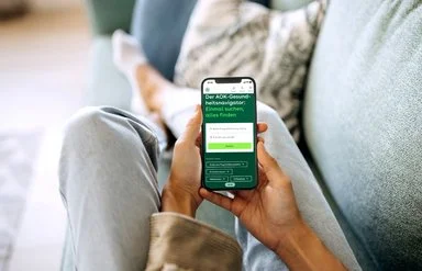 Eine nicht erkennbare Person sitzt auf einem Sofa und hält ein Smartphone in den Händen, im Display ist der AOK-Gesundheitsnavigator zu sehen.