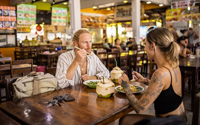 Ein Mann und eine Frau essen mit Stäbchen in einem Restaurant. Auf dem Tisch stehen neben den Tellern zwei aufgeschnittene grüne Kokosnüsse, in denen sich Strohhalme befinden.