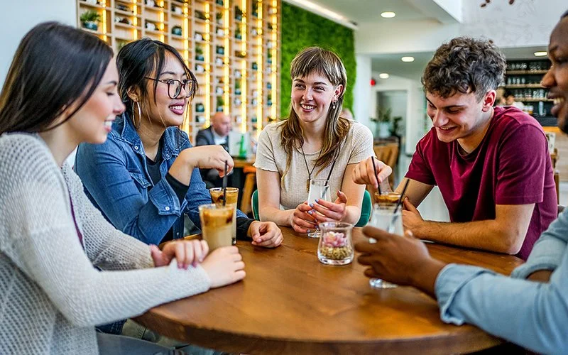 Eine Gruppe von vier jungen Freundinnen und Freunden sitzt an einem Tisch in einem Café und teilt einen fröhlichen, gemeinsamen Moment.