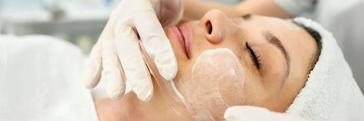 Zu sehen ist das Gesicht einer Frau, die Haare und den Oberkörper mit einem weißen Handtuch abgedeckt. Die Frau hat ihre Augen geschlossen und macht einen entspannten Eindruck. Eine andere, nicht auf dem Bild sichtbare Person streicht ihr mit weißen Schutzhandschuhen eine weißliche Creme ins Gesicht.