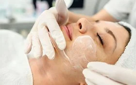 Zu sehen ist das Gesicht einer Frau, die Haare und den Oberkörper mit einem weißen Handtuch abgedeckt. Die Frau hat ihre Augen geschlossen und macht einen entspannten Eindruck. Eine andere, nicht auf dem Bild sichtbare Person streicht ihr mit weißen Schutzhandschuhen eine weißliche Creme ins Gesicht.