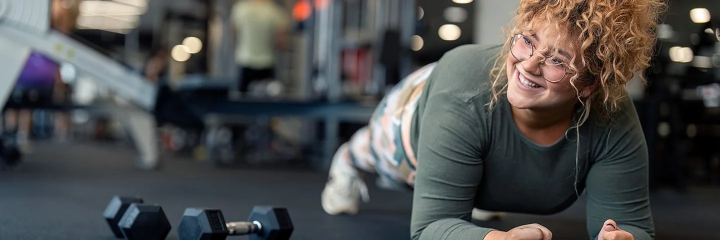 Lächelnde Frau mit lockigen Haaren macht im Fitnessstudio eine Unterarmstütz-Übung auf einer Trainingsmatte, neben ihr liegen zwei Kurzhanteln.