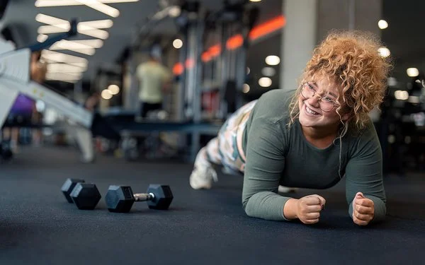 Lächelnde Frau mit lockigen Haaren macht im Fitnessstudio eine Unterarmstütz-Übung auf einer Trainingsmatte, neben ihr liegen zwei Kurzhanteln.