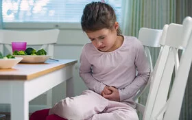 Ein Mädchen sitzt am Küchentisch und hält sich mit schmerzverzerrtem Gesicht den Bauch – vielleicht steckt eine Lebensmittelallergie dahinter.