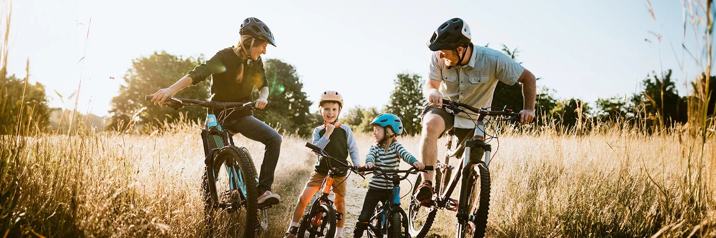 Eine Familie, bestehend aus Vater, Mutter und zwei kleinen Jungen, auf Fahrrädern, die eine Pause auf einem Feldweg einlegen. Alle tragen Helme und lächeln sich gegenseitig fröhlich an.