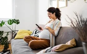 Eine schwangere Frau sitzt im Wohnzimmer auf dem Sofa und schaut auf ihr Handy.