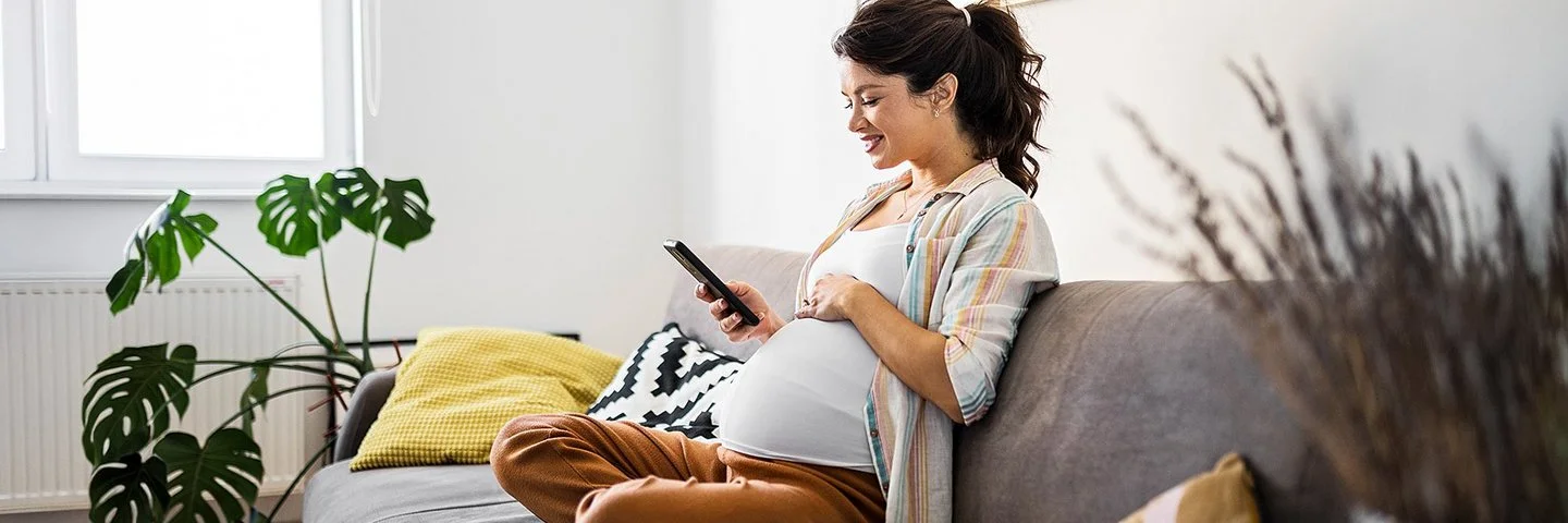 Eine schwangere Frau sitzt im Wohnzimmer auf dem Sofa und schaut auf ihr Handy.