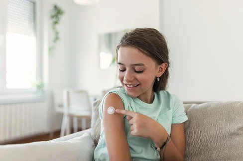 Ein junges Mädchen sitzt auf dem Sofa und zeigt auf kleines Pflaster an ihrem Oberarm.