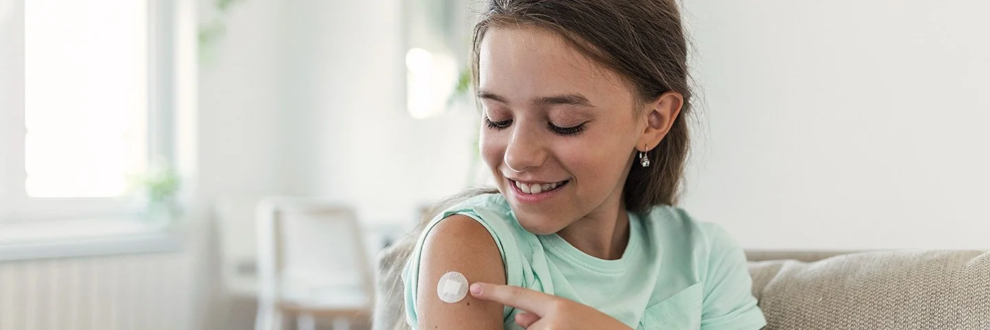 Ein junges Mädchen sitzt auf dem Sofa und zeigt auf kleines Pflaster an ihrem Oberarm.
