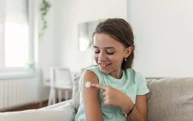 Ein junges Mädchen sitzt auf dem Sofa und zeigt auf kleines Pflaster an ihrem Oberarm.