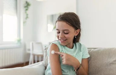 Ein junges Mädchen sitzt auf dem Sofa und zeigt auf kleines Pflaster an ihrem Oberarm.