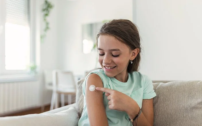 Ein junges Mädchen sitzt auf dem Sofa und zeigt auf kleines Pflaster an ihrem Oberarm.
