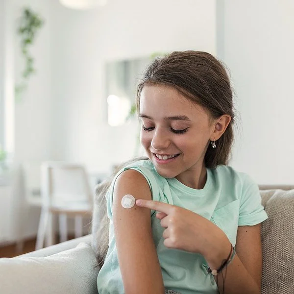 Ein junges Mädchen sitzt auf dem Sofa und zeigt auf kleines Pflaster an ihrem Oberarm.