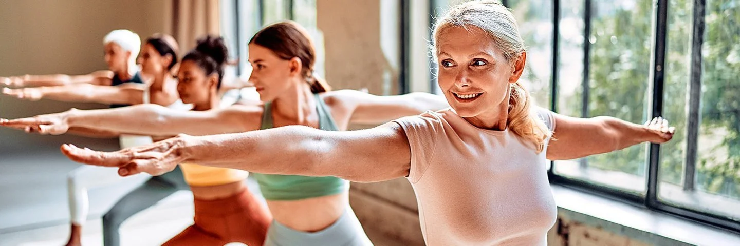 Fünf Frauen in sportlicher Kleidung praktizieren in einem Yoga-Studio auf Matten die Yoga-Übung „Krieger 2“.