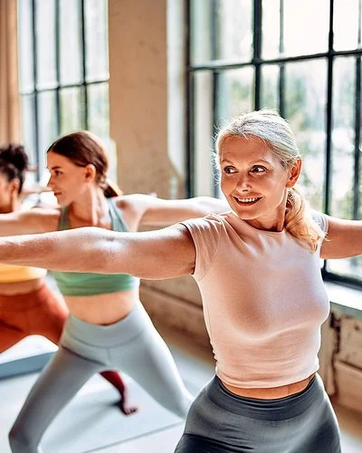 Fünf Frauen in sportlicher Kleidung praktizieren in einem Yoga-Studio auf Matten die Yoga-Übung „Krieger 2“.