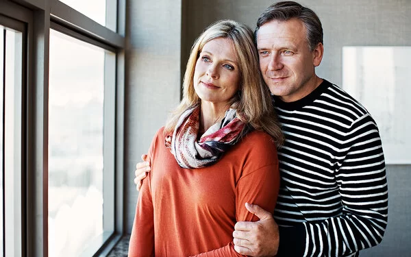 Die Partnerin mit COPD unterstützen: Ein Mann umarmt von hinten seine Frau, gemeinsam schauen sie aus einem Fenster.