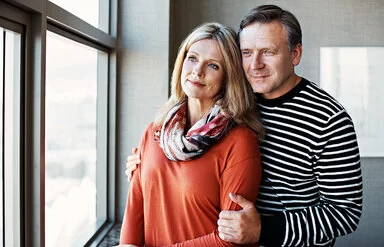 Die Partnerin mit COPD unterstützen: Ein Mann umarmt von hinten seine Frau, gemeinsam schauen sie aus einem Fenster.