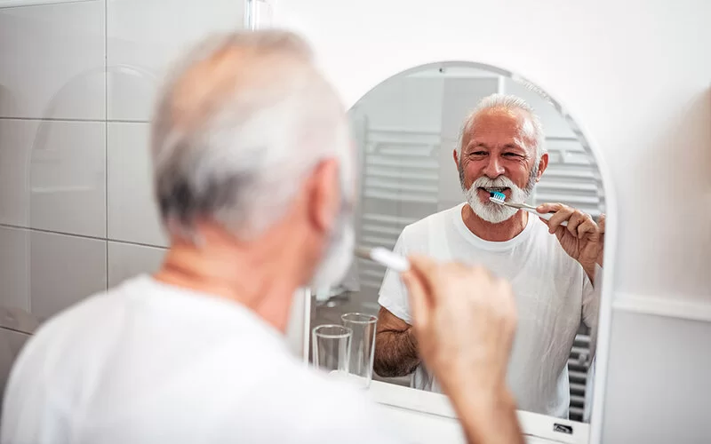 Älterer Mann steht im Badezimmer am Becken und putzt sich seine Zähne.