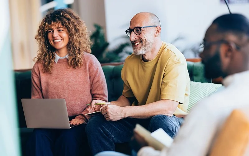 Eine Frau und zwei Männer sitzen mit Laptops und Notizbüchern auf einer Couch und lachen.