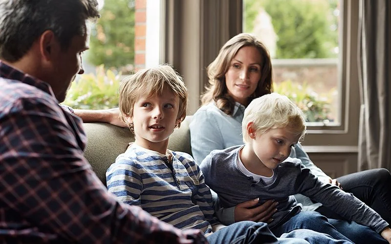 Eine vierköpfige Familie sitzt gemeinsam auf einem Sofa einer häuslichen Umgebung und unterhält sich.