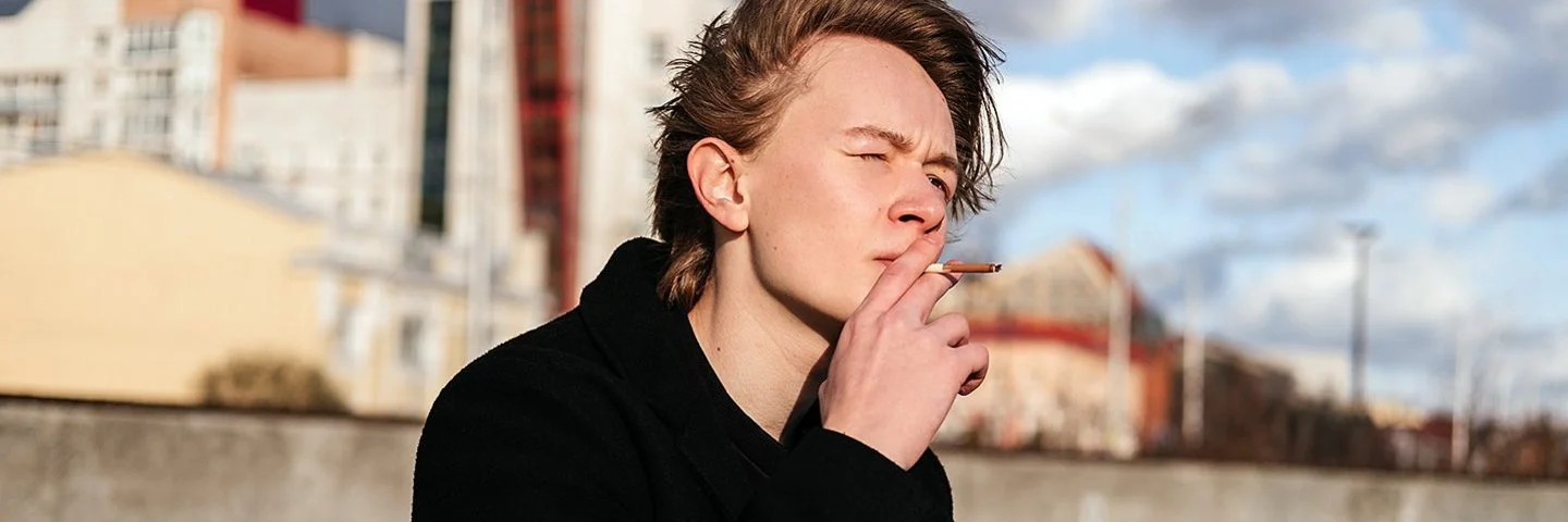 Ein Teenager sitzt vor der Skyline einer Stadt und raucht eine Zigarette.