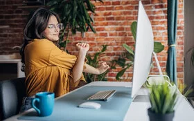 Eine junge Frau mit Brille und senfgelber Bluse sitzt an einem Schreibtisch vor einem Computer und streckt sich.