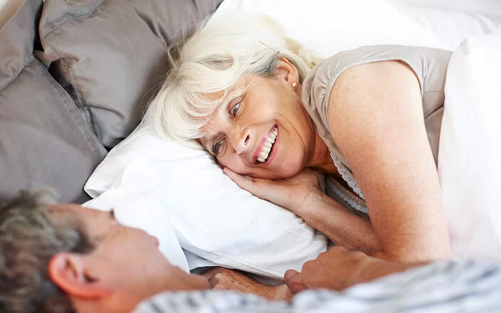 Ein Mann und eine Frau, beide leicht ergraut, liegen in Schlafkleidung im Bett und blicken sich lächelnd an.