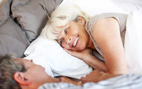 Ein Mann und eine Frau, beide leicht ergraut, liegen in Schlafkleidung im Bett und blicken sich lächelnd an.