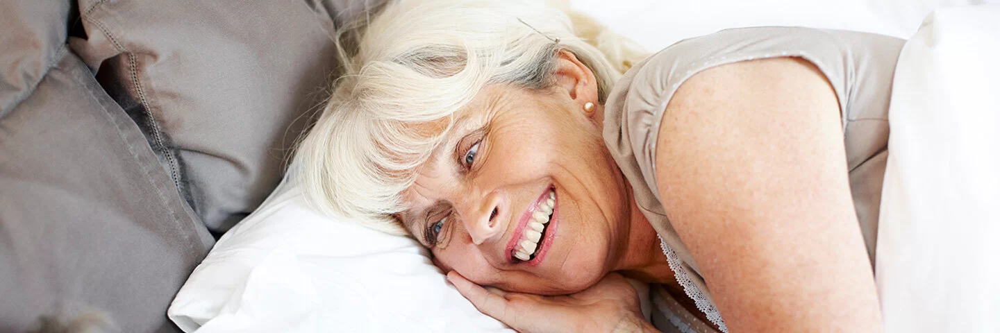 Ein Mann und eine Frau, beide leicht ergraut, liegen in Schlafkleidung im Bett und blicken sich lächelnd an.