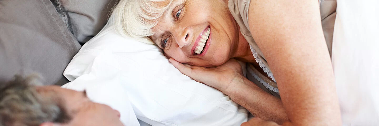 Ein Mann und eine Frau, beide leicht ergraut, liegen in Schlafkleidung im Bett und blicken sich lächelnd an.