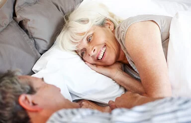 Ein Mann und eine Frau, beide leicht ergraut, liegen in Schlafkleidung im Bett und blicken sich lächelnd an.
