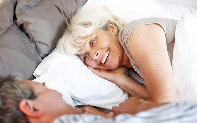 Ein Mann und eine Frau, beide leicht ergraut, liegen in Schlafkleidung im Bett und blicken sich lächelnd an.
