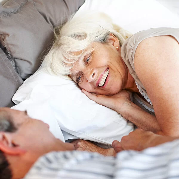 Ein Mann und eine Frau, beide leicht ergraut, liegen in Schlafkleidung im Bett und blicken sich lächelnd an.