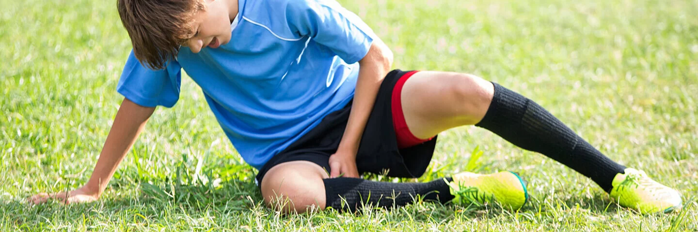 Ein Junge in Fußballkleidung sitzt mit Schmerzen auf dem Rasen und fasst sich an den Oberschenkel.