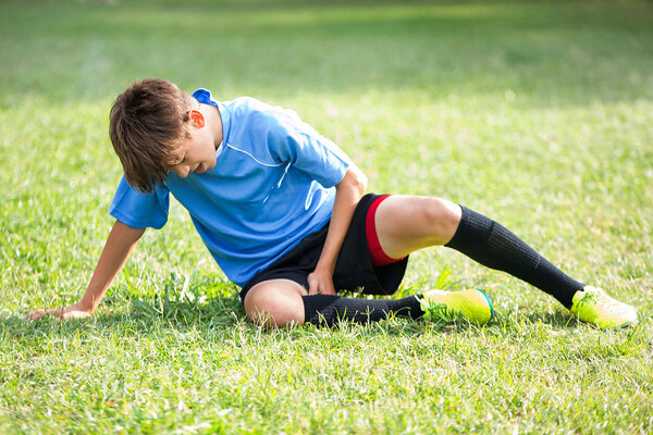 Ein Junge in Fußballkleidung sitzt mit Schmerzen auf dem Rasen und fasst sich an den Oberschenkel.
