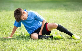 Ein Junge in Fußballkleidung sitzt mit Schmerzen auf dem Rasen und fasst sich an den Oberschenkel.