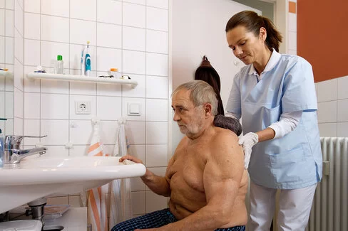 Eine Pflegeperson wäscht einem älteren Mann den Rücken. Die Frau trägt Einmalhandschuhe, die als zum Verbrauch bestimmte Pflegehilfsmittel gelten.