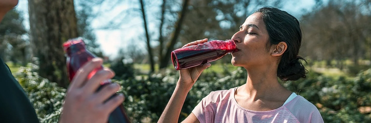 Ein sportlich gekleidetes Paar in einem Park, beide trinken Rote Bete-Saft aus Flaschen.