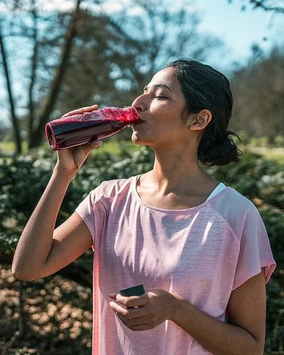 Ein sportlich gekleidetes Paar in einem Park, beide trinken Rote Bete-Saft aus Flaschen.