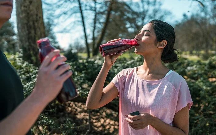 Ein sportlich gekleidetes Paar in einem Park, beide trinken Rote Bete-Saft aus Flaschen.