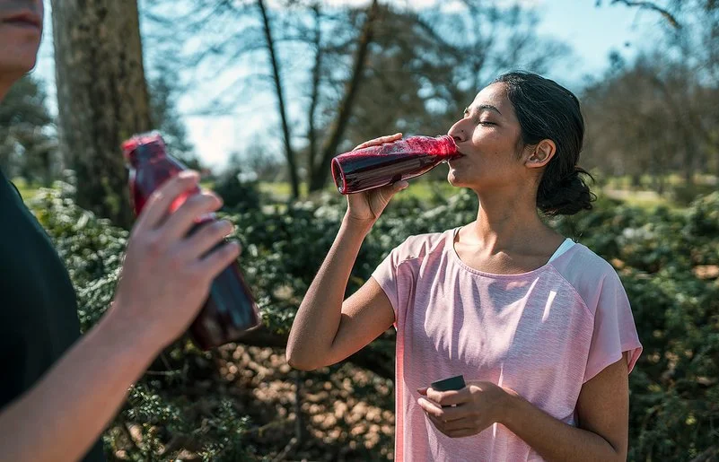 Ein sportlich gekleidetes Paar in einem Park, beide trinken Rote Bete-Saft aus Flaschen.
