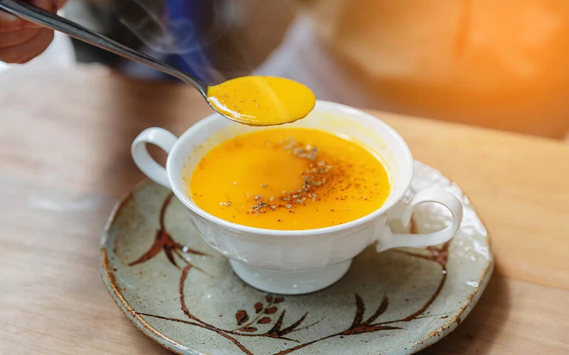 Auf einem Tisch steht eine kleine Suppentasse mit Kürbissuppe. Eine Hand hält einen mit Suppe gefüllten Löffel über der Tasse.
