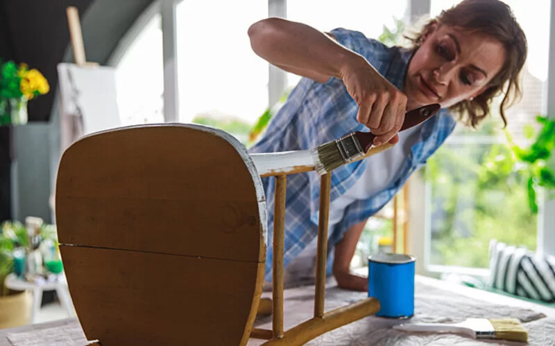 Eine Frau betreibt Upcycling indem sie einen alten Holzstuhl mit weißer Farbe restauriert.