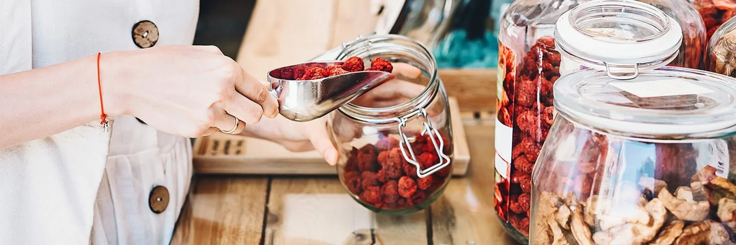 Nahaufnahme einer Person, die in einem Unverpackt-Laden mit einer Metallschaufel getrocknete rote Beeren in ein Glas füllt.