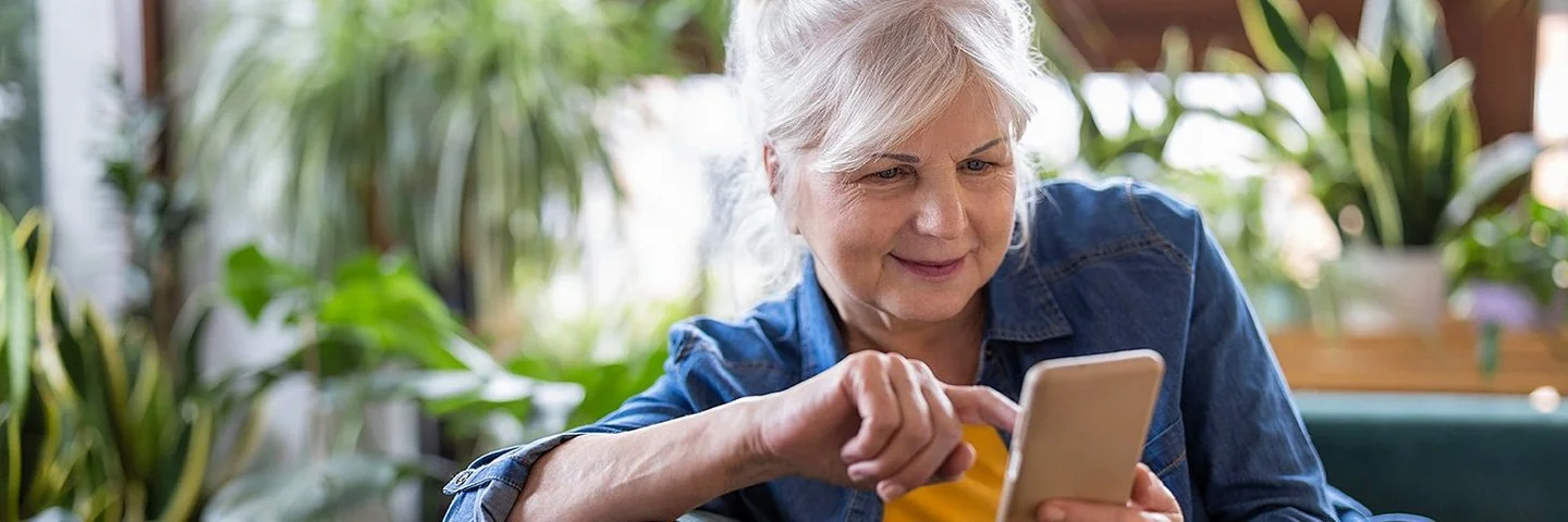 Eine ältere Frau sitzt entspannt auf dem Sofa und tippt etwas in ihr Smartphone ein.