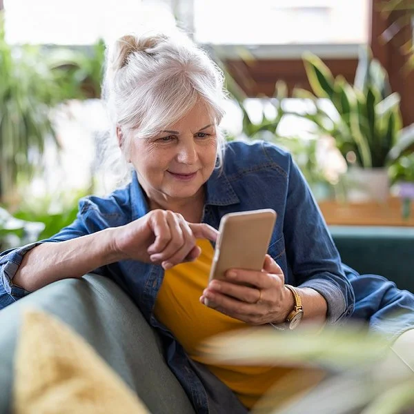 Eine ältere Frau sitzt entspannt auf dem Sofa und tippt etwas in ihr Smartphone ein.