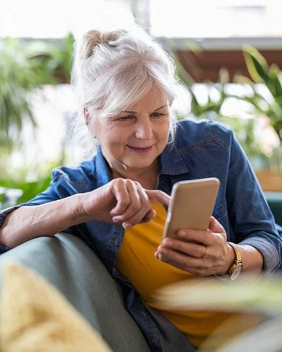 Eine ältere Frau sitzt entspannt auf dem Sofa und tippt etwas in ihr Smartphone ein.