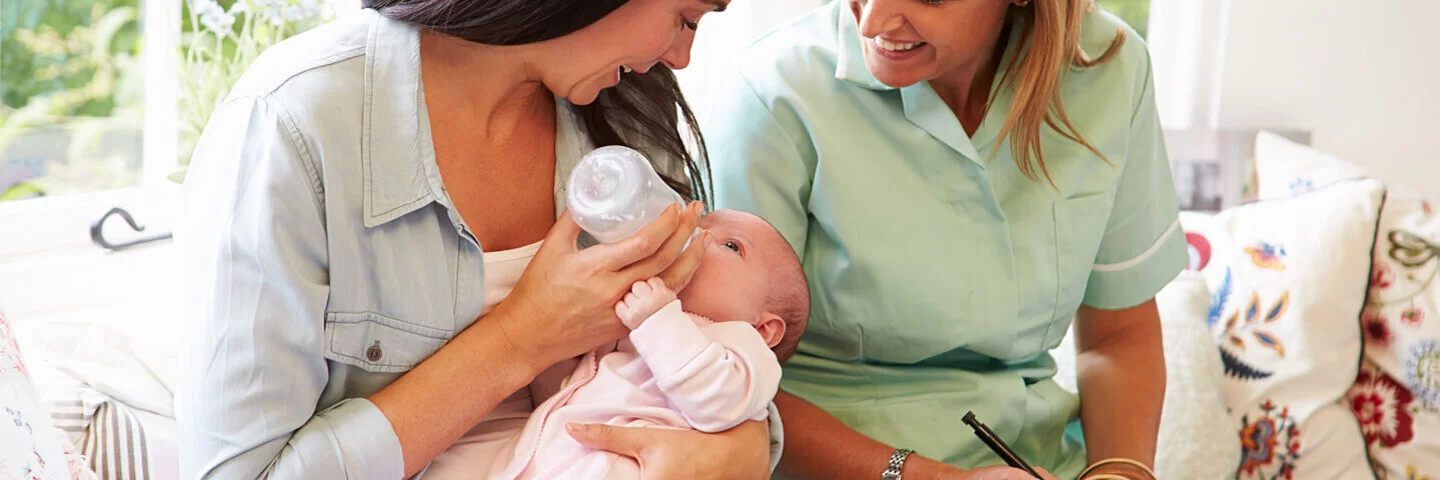 Hebamme unterstützt junge Mutter beim Fläschchengeben ihres Babys.